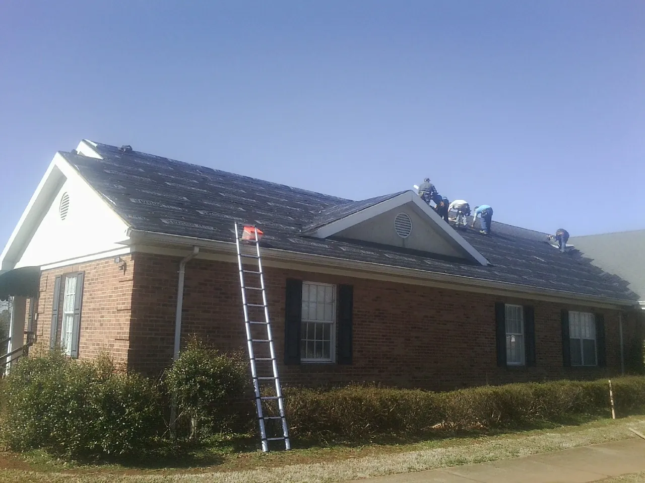 Crew tearing shingles off a brick ranch roof with a ladder set — Emergency Roof Repair work by our crew in Essex Junction