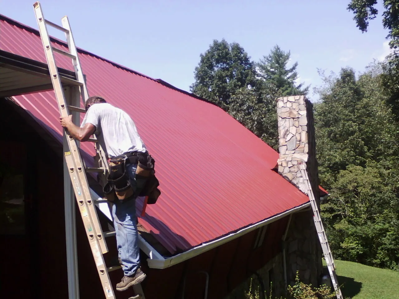 Roofer on a ladder beside a red metal roof and stone chimney — Soffit & Fascia Repair by Roofing Brothers in Essex Junction